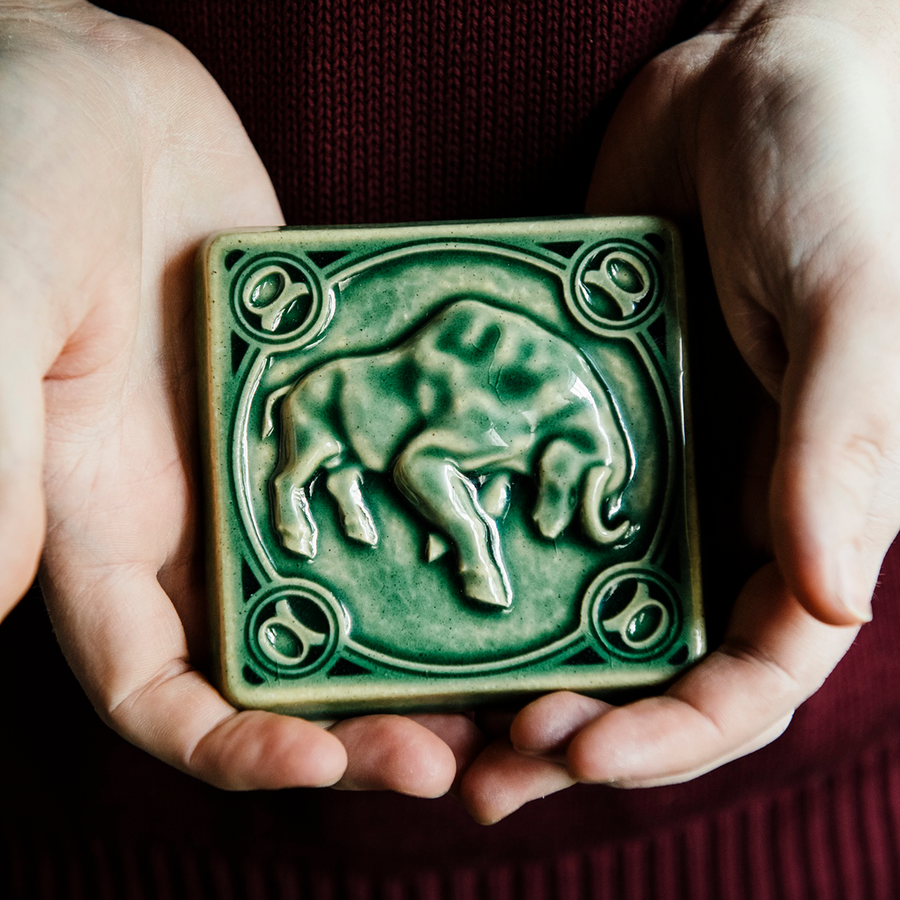 The glossy green Emerald glaze pools across the low-relief sculpted bull form of this Taurus tile. Two graceful hands hold the tile in natural light to show off the glossy surface.