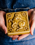 The glossy gold Honey Gloss glaze pools across the low-relief sculpted ram form of this Aries tile. Two graceful hands hold the tile in natural light to show off the glossy surface.
