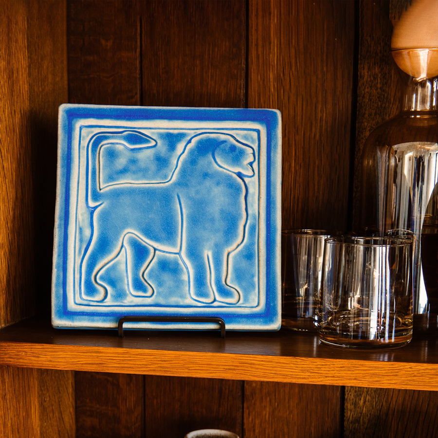 This Aztec Lion tile features the matte bright blue Sky glaze. The lion faces to the right and rests on the wooden bookshelf of a collector.