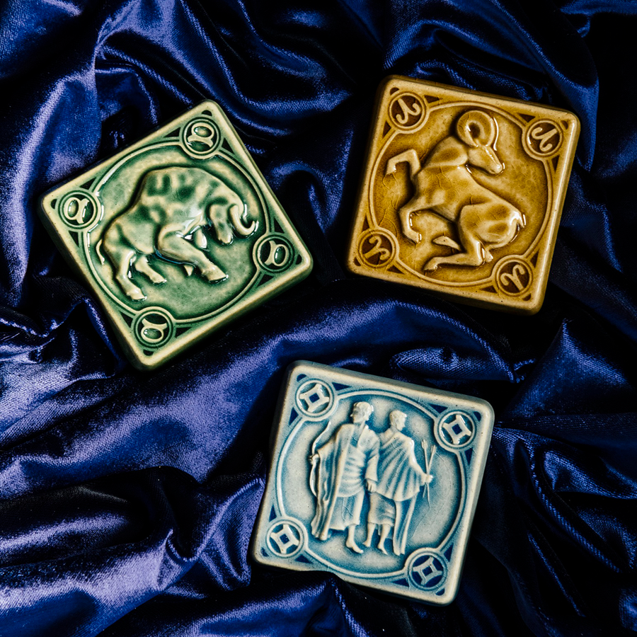 The Gemini tile lies with the other two Pewabic Zodiac tiles on rich, velvety fabric the color of the night sky. The Taurus tile features a sturdy bull with its head bowed and horns lowered. The Aries tile has an alert ram with its legs positioned as if it is prancing. The ram's head is turned backwards over its shoulder.