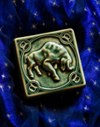 The center of this 4 inch by 4 inch Taurus tile features a sturdy bull with its head bowed and horns lowered. The border around the square tile encircles the bull and each of the four corners contains a circle with the Taurus sign in the center. The tile rests on a deep blue velvet fabric with gold stars speckled across its surface.