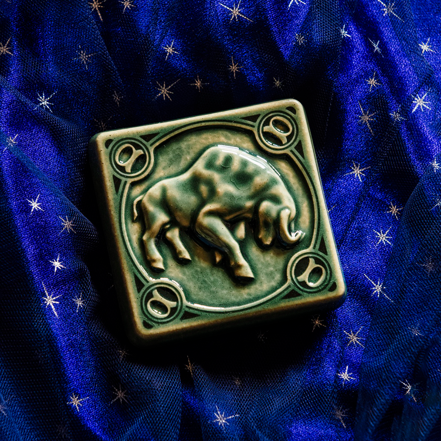 The center of this 4 inch by 4 inch Taurus tile features a sturdy bull with its head bowed and horns lowered. The border around the square tile encircles the bull and each of the four corners contains a circle with the Taurus sign in the center. The tile rests on a deep blue velvet fabric with gold stars speckled across its surface.