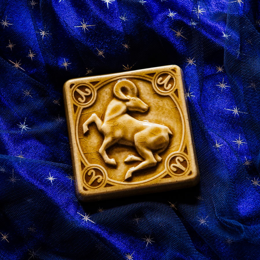 The center of this 4 inch by 4 inch Aries tile features an alert ram with its legs positioned as if it is prancing. The ram's head is turned backwards over its shoulder. The border around the square tile encircles the beast and each of the four corners contains the Aries sign. The tile rests on a deep blue velvet fabric with gold stars speckled across its surface.