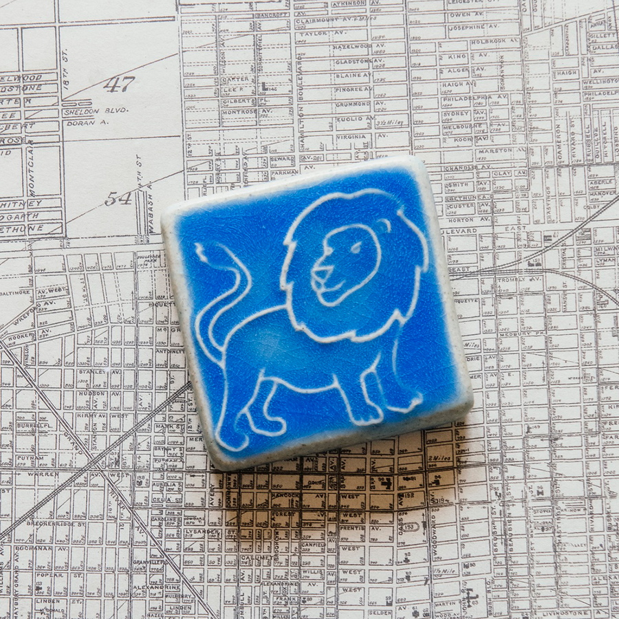 The three inch by three inch Lion tile features a hand-drawn cartoonish lion with a scraggly mane and swishing tail. The tile rests on a black and white map of Detroit.