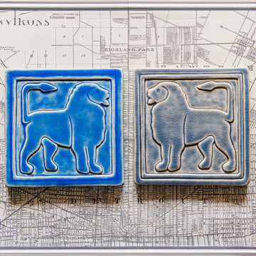 Two 8 inch by 8 inch tiles sit on a black and white map of Detroit. Each tile features a simplified silhouette of a lion's body, each of their tails are alert and pointed forward to their head. The tiles are mirrored, with the bright blue tile on the left facing the gray-blue tile on the right. 