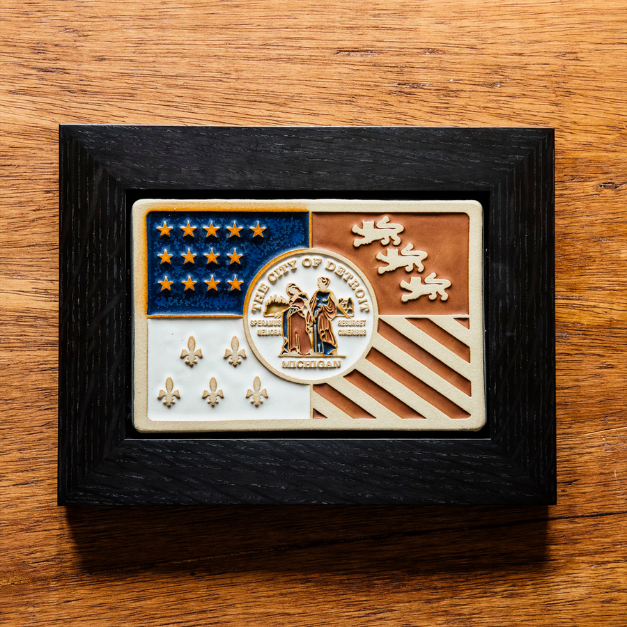 Our framed, hand-painted Detroit Flag Tile is laying on the historic Pewabic tiled floor in our upstairs gallery space. These tiles are a rustic brown and tawny in appearance.