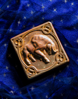 The center of this 4 inch by 4 inch Taurus tile features a sturdy bull with its head bowed and horns lowered. The border around the square tile encircles the bull and each of the four corners contains a circle with the Taurus sign in the center. The tile rests on a deep blue velvet fabric with gold stars speckled across its surface.