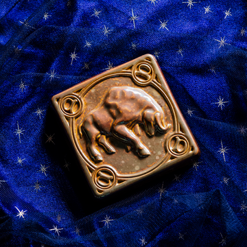 The center of this 4 inch by 4 inch Taurus tile features a sturdy bull with its head bowed and horns lowered. The border around the square tile encircles the bull and each of the four corners contains a circle with the Taurus sign in the center. The tile rests on a deep blue velvet fabric with gold stars speckled across its surface.