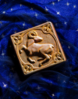 The center of this 4 inch by 4 inch Aries tile features an alert ram with its legs positioned as if it is prancing. The ram's head is turned backwards over its shoulder. The border around the square tile encircles the beast and each of the four corners contains the Aries sign. The tile rests on a deep blue velvet fabric with gold stars speckled across its surface.