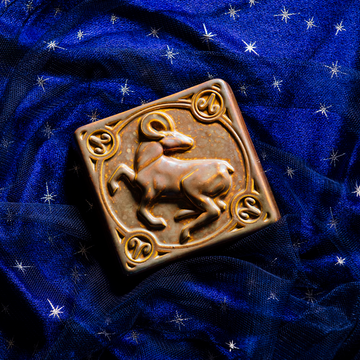 The center of this 4 inch by 4 inch Aries tile features an alert ram with its legs positioned as if it is prancing. The ram's head is turned backwards over its shoulder. The border around the square tile encircles the beast and each of the four corners contains the Aries sign. The tile rests on a deep blue velvet fabric with gold stars speckled across its surface.