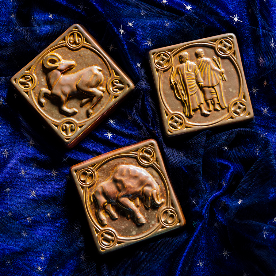 The Gemini tile lies with the other two Pewabic Zodiac tiles on rich, velvety fabric the color of the night sky. The Taurus tile features a sturdy bull with its head bowed and horns lowered. The Aries tile has an alert ram with its legs positioned as if it is prancing. The ram's head is turned backwards over its shoulder.
