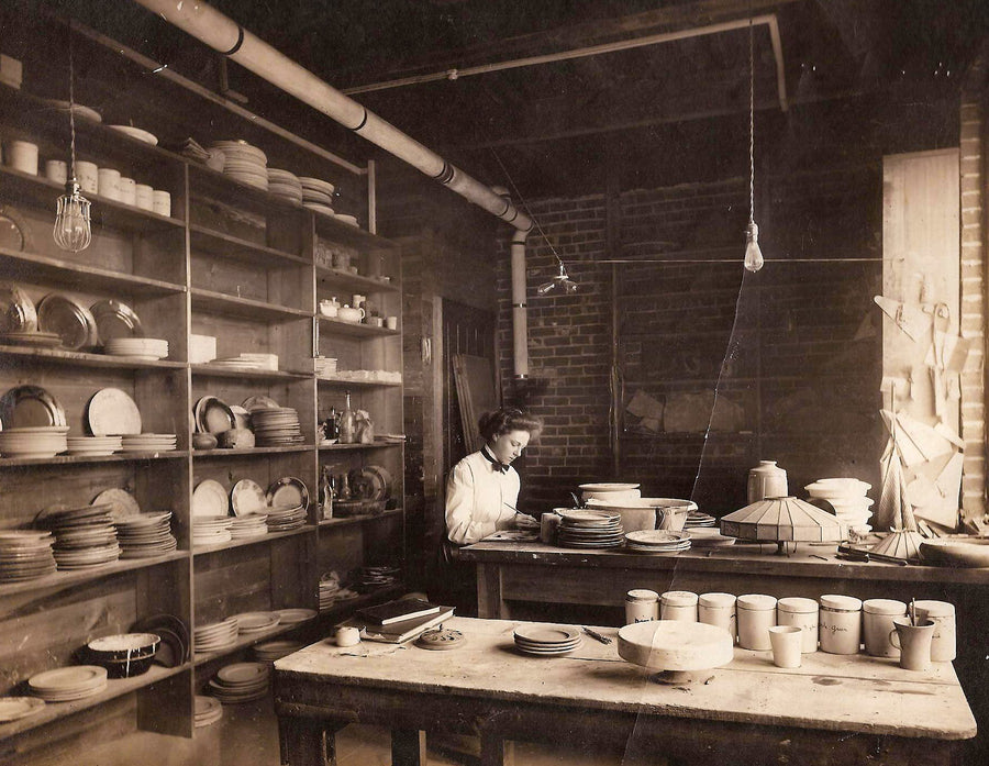 A historic image of Pewabic co-founder Mary Chase Perry Stratton. She sits at a work table in a room full of plates, bowls and other vessels.