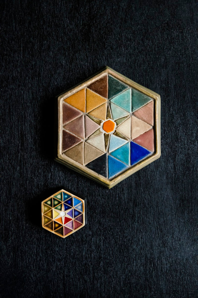 A modern Hex tile is displayed next to its historic counterpart. Both hexagonal tiles feature a geometric pattern with each small section filled with a different colorful glaze. These tiles are used to showcase all of the colors in Pewabic's glaze palette.