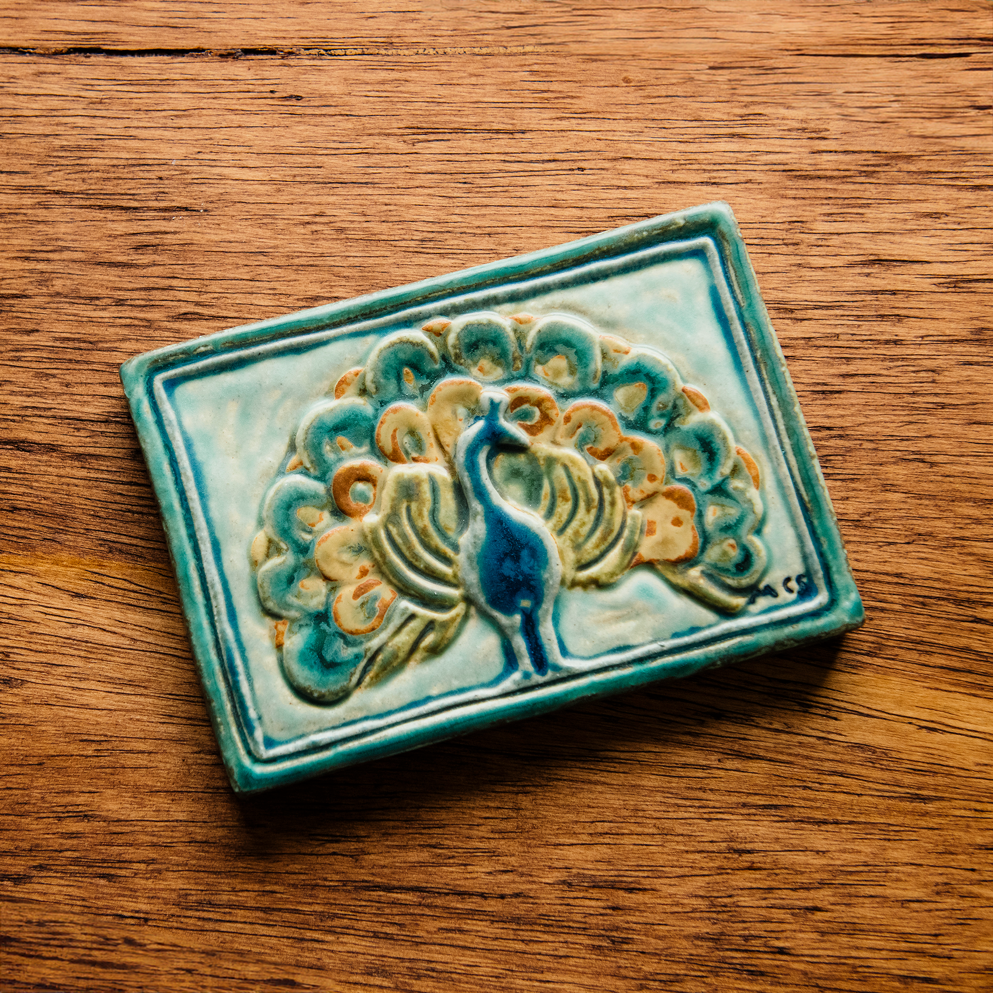 The Peacock Tile features a peacock facing straight ahead with its feathers fanned out showing off its colorful plumes. In the bottom right corner of the tile are the letters "M C S" in black writing. This is the only tile on which Pewabic's co-founder Mary Chase Stratton included a signature. The hand painted tile features bright blues, greens and beiges.