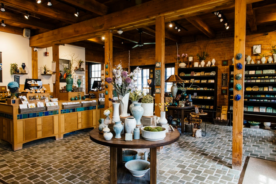 A view of our Gallery store in the historic Pewabic building. Colorful vases sit on a round wooden table. Shelves full of tiles in all shapes and sizes line the brick walls. Wooden beams and a decorative tile floor decorate the space. 