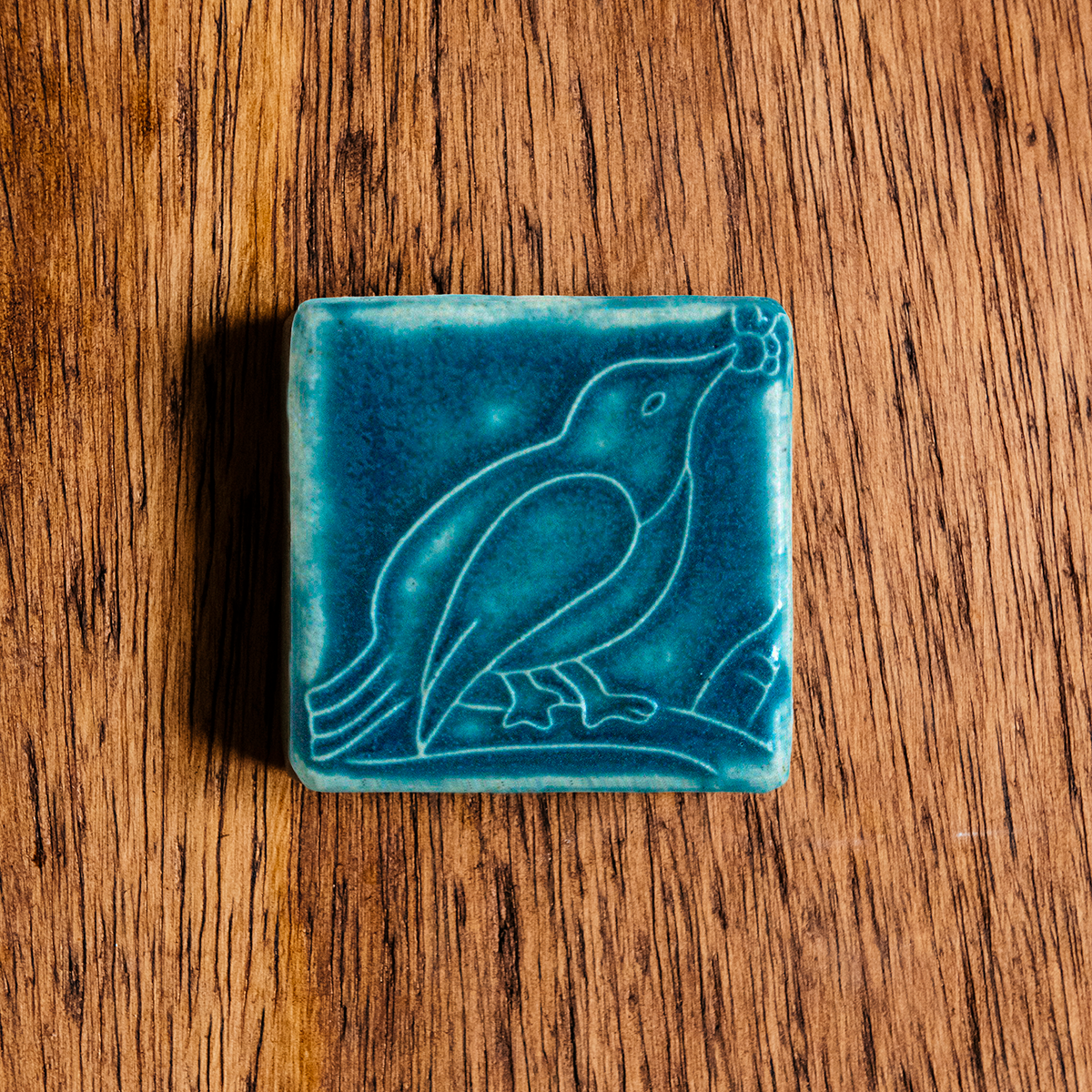 The ceramic Bird with Berries Tile depicts a bird sitting on a branch with berries in its beak. This tile is glazed in a matte Peacock blue color.