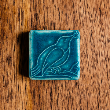 The ceramic Bird with Berries Tile depicts a bird sitting on a branch with berries in its beak. This tile is glazed in a matte Peacock blue color.