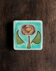 The Hand-Painted Rose Tile features a line drawing of a pinkish dusty rose in the center with a green stem and leaves on a pale blue background.