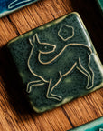 This Prancing Deer Tile features a line drawing of a female deer. Her legs are lifted off the ground in a dancing motion while her head is turned toward her back. A small flower is floating above her body.