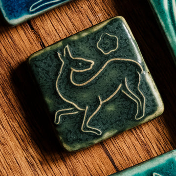 This Prancing Deer Tile features a line drawing of a female deer. Her legs are lifted off the ground in a dancing motion while her head is turned toward her back. A small flower is floating above her body.