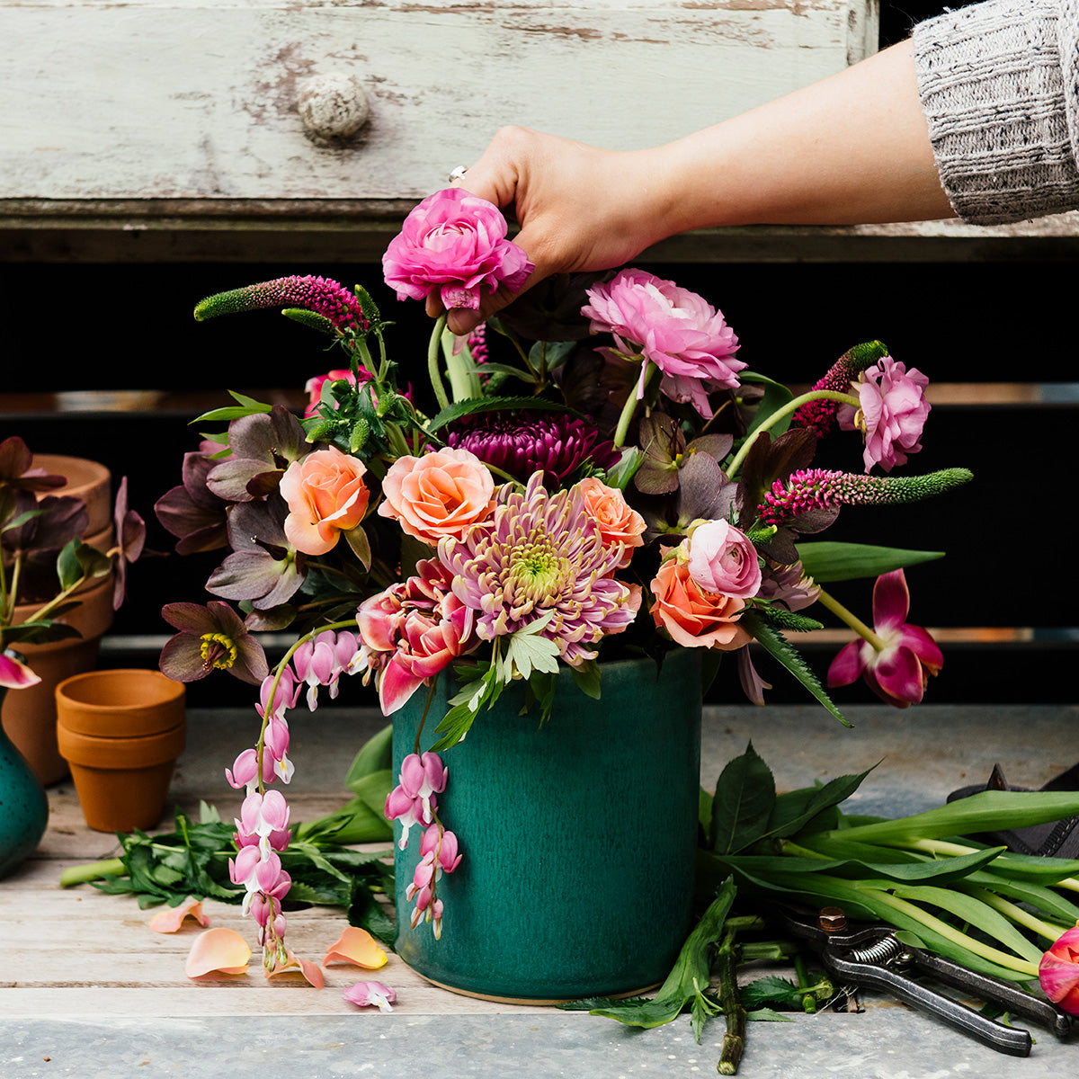 This Pewabic Blue crock sits outside on a wooden work table covered in petals and garden snips. A hand reaches into frame to place a flower, just plucked from the garden into the mostly finished bouquet. 