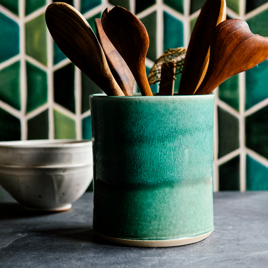 This Pewabic green Crock is holding wooden cooking utensils on a kitchen counter. The backsplash features multicolored green Pewabic tiles in a hexagonal design.