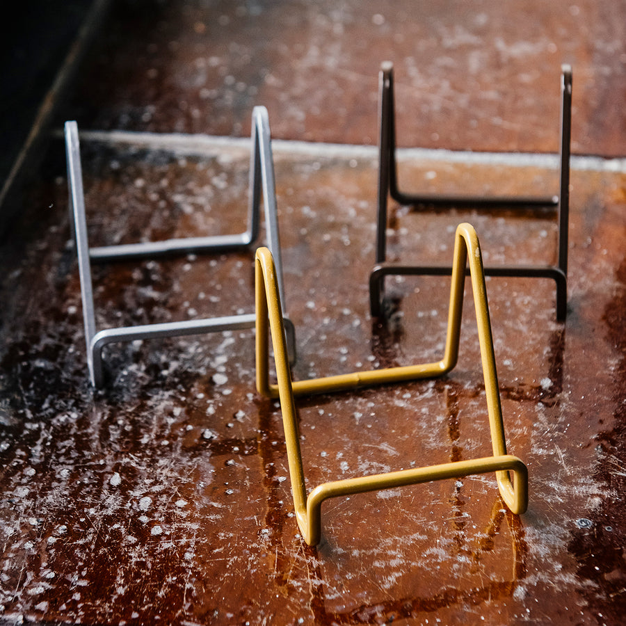 All three matte gold, silver, and bronze tile stands photographed together on a tiled-window ledge.