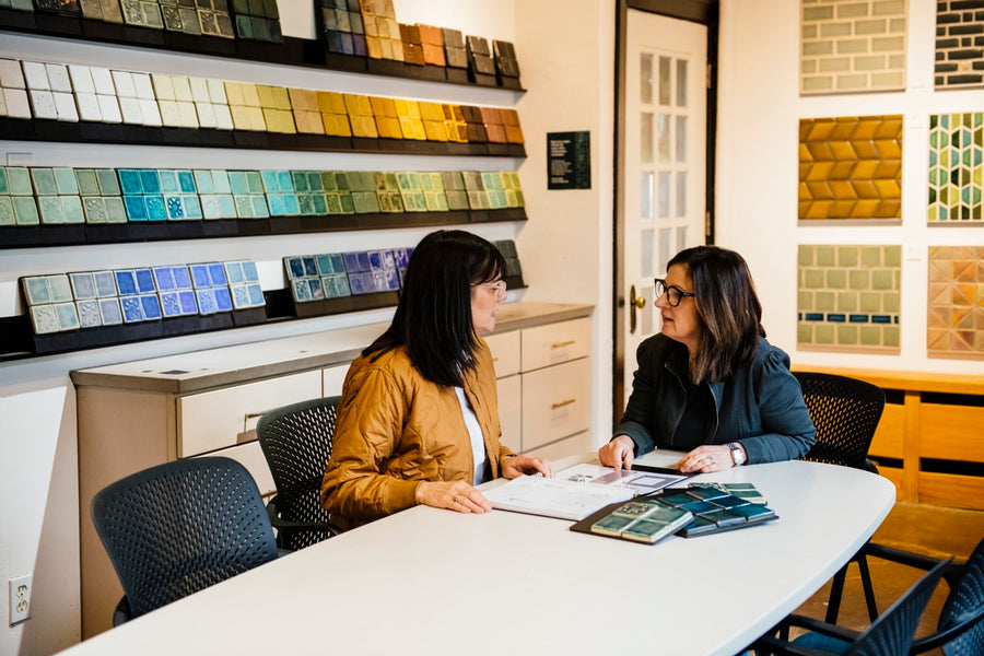 A Pewabic designer sits at a table in our tile showroom with a client. They look over a portfolio folder with colorful tile samples strewn around them.