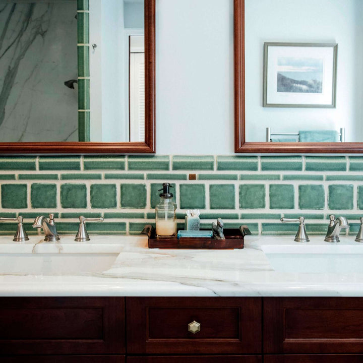 Green Pewabic tiles in square and rectangular shapes line the wall behind a counter with double sinks in a bathroom.