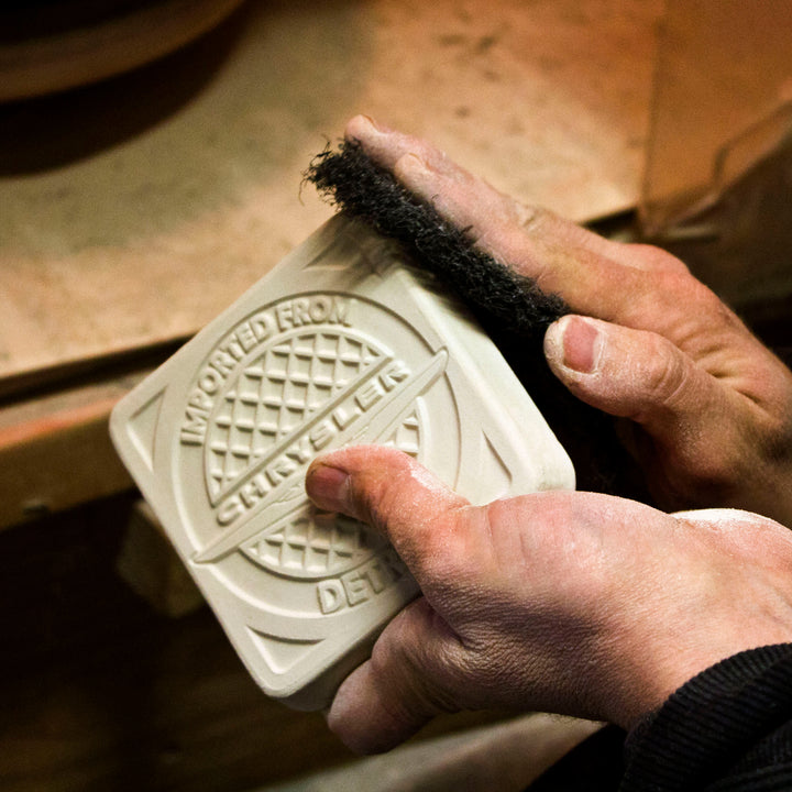 A Pewabic artisan shaves down the sides of an unfinished commemorative tile. The tile is embossed with a Chrysler logo and the words "Imported from Detroit" surround the illustration.