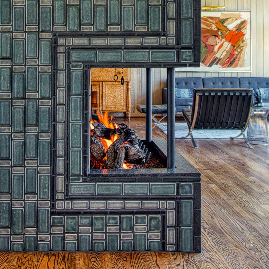 Side view of the lit fireplace. Two mid-century modern chairs and a cozy living room area are visible in the frame. The natural-finished wood vaulted ceiling is a beautiful contrast to the strong geometric design of the fireplace installation.