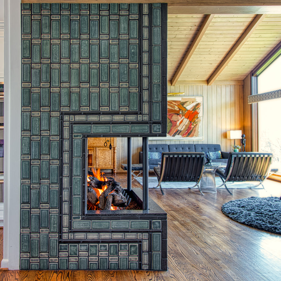 Side view of the lit fireplace. Two mid-century modern chairs and a cozy living room area are visible in the frame. The natural-finished wood vaulted ceiling is a beautiful contrast to the strong geometric design of the fireplace installation.