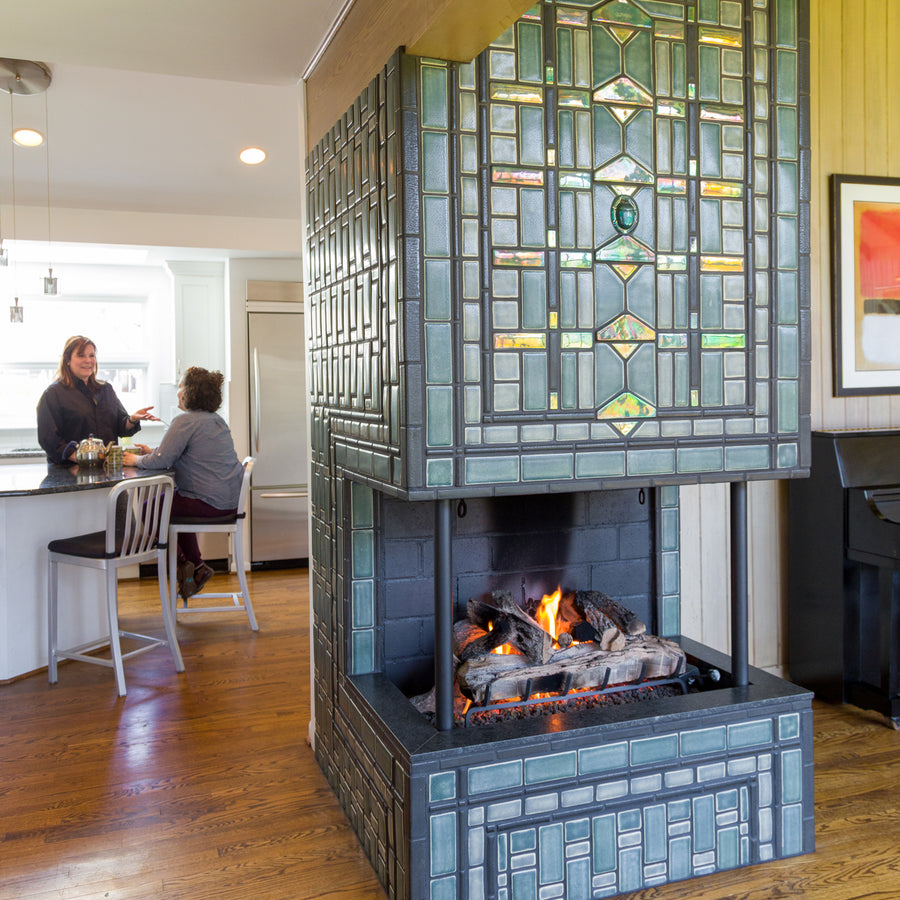 Two women converse at a kitchen island over a cup of tea. The custom three-sided fireplace is in view. From this perspective, the Iridescent tile details and featured Scarab motif are very visible. The fire is lit, creating a cozy and inviting atmosphere. 