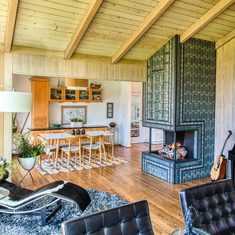 A quarter view of a three-sided custom Pewabic Tile fireplace with a strong geometric pattern of subway and custom-cut tiles. The client’s furniture, including their dining table and cow-hide lounge chair are visible in the frame. 