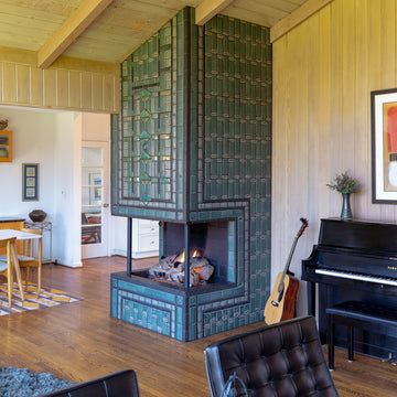 A quarter view of a three-sided custom Pewabic Tile fireplace with a strong geometric pattern of subway and custom-cut tiles. The front of the fireplace prominently incorporates our signature Iridescent Aurora glaze with a Matte Green Iridescent Scarab Paperweight at the center. There is a wall piano in the corner and the fireplace is set in a spacious mid-century modern ranch home. 