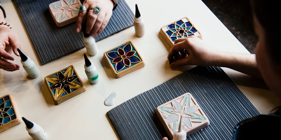 Attendees at an interactive Tile Glazing Workshop. They are in various stages of hand-painting their selected tile design. Finished examples run the length of the table in front of them.