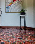 A tiled foyer comprised of warm, red and earth toned tiles with bright, luminous "blush" iridescent tiles that look golden with the sunshine creeping in. There is a large mirror-like charcoal glazed Pewabic vase with a plant on a stand in-frame against thew wall. There is a colorful, abstract painting of grays, blues, reds, and tans above and to the left of the vase.