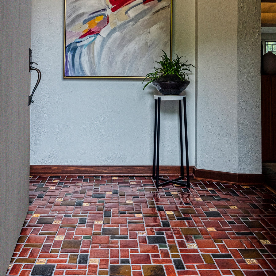 A tiled foyer comprised of warm, red and earth toned tiles with bright, luminous "blush" iridescent tiles that look golden with the sunshine creeping in. There is a large mirror-like charcoal glazed Pewabic vase with a plant on a stand in-frame against thew wall. There is a colorful, abstract painting of grays, blues, reds, and tans above and to the left of the vase.