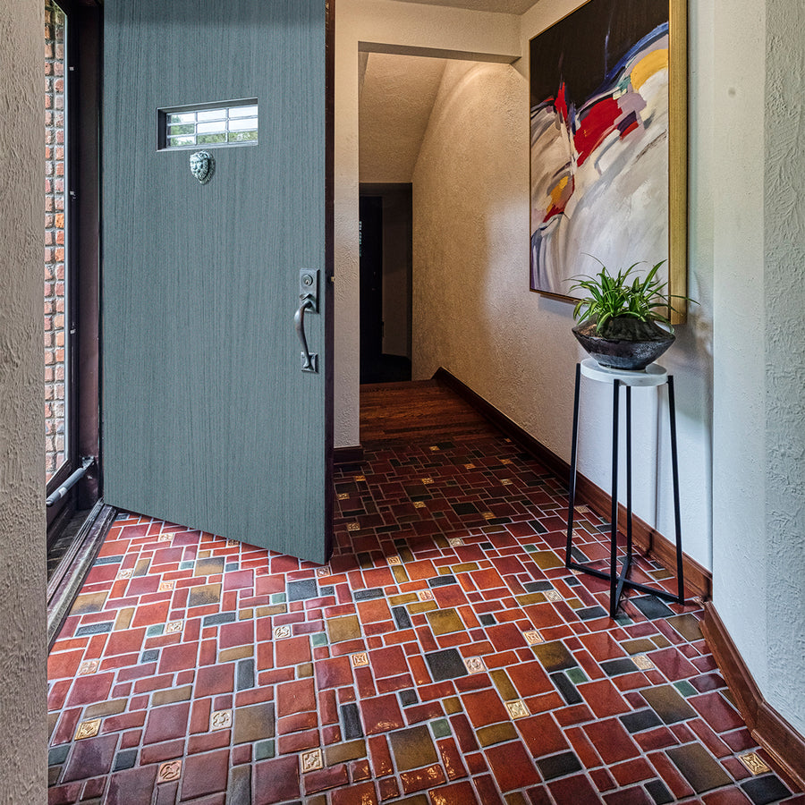 A door opens up to a tiled foyer comprised of warm, red and earth toned tiles with bright, luminous "blush" iridescent tiles that look golden with the sunshine creeping in. There is a large mirror-like charcoal glazed Pewabic vase with a plant on a stand in-frame against thew wall. The photo is taken at an angle to the right of the open door.