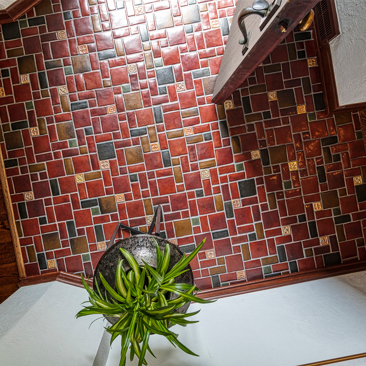 A door opens up to a tiled foyer comprised of warm, red and earth toned tiles with bright, luminous "blush" iridescent tiles that look golden with the sunshine creeping in. There is a large mirror-like charcoal glazed Pewabic vase with a plant on a stand in-frame against thew wall. The photo is taken from an "aerial" vantage point.