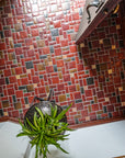 A door opens up to a tiled foyer comprised of warm, red and earth toned tiles with bright, luminous "blush" iridescent tiles that look golden with the sunshine creeping in. There is a large mirror-like charcoal glazed Pewabic vase with a plant on a stand in-frame against thew wall. The photo is taken from an "aerial" vantage point.