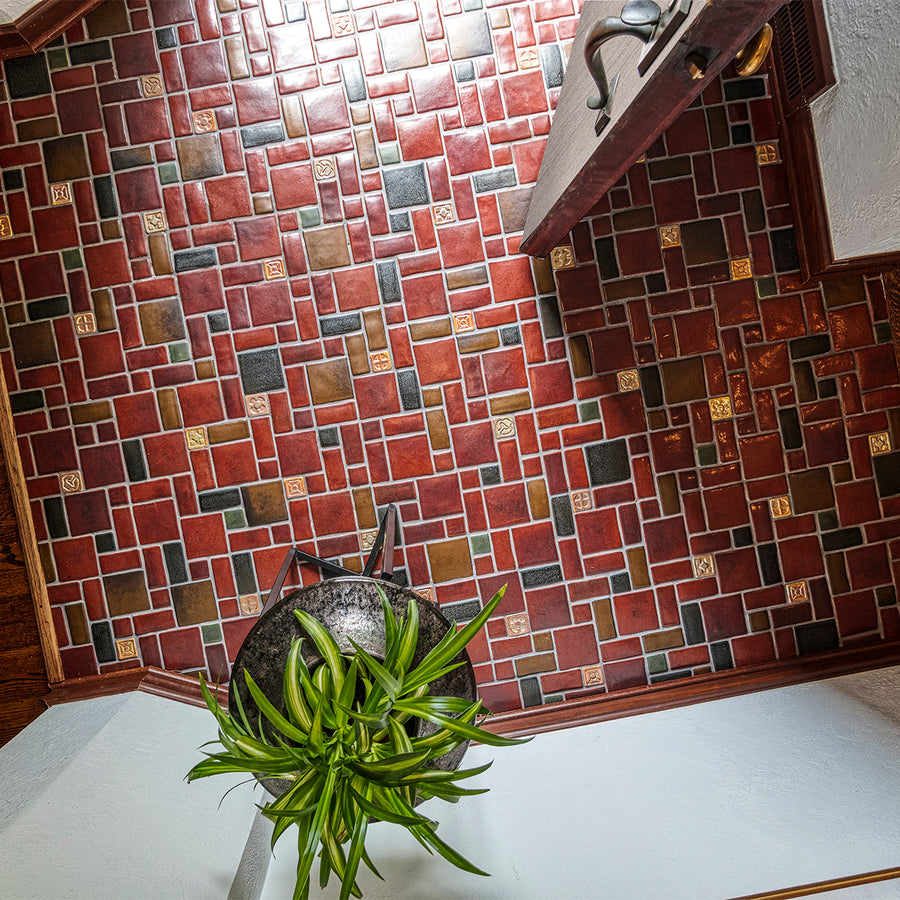 A door opens up to a tiled foyer comprised of warm, red and earth toned tiles with bright, luminous "blush" iridescent tiles that look golden with the sunshine creeping in. There is a large mirror-like charcoal glazed Pewabic vase with a plant on a stand in-frame against thew wall. The photo is taken from an "aerial" vantage point.