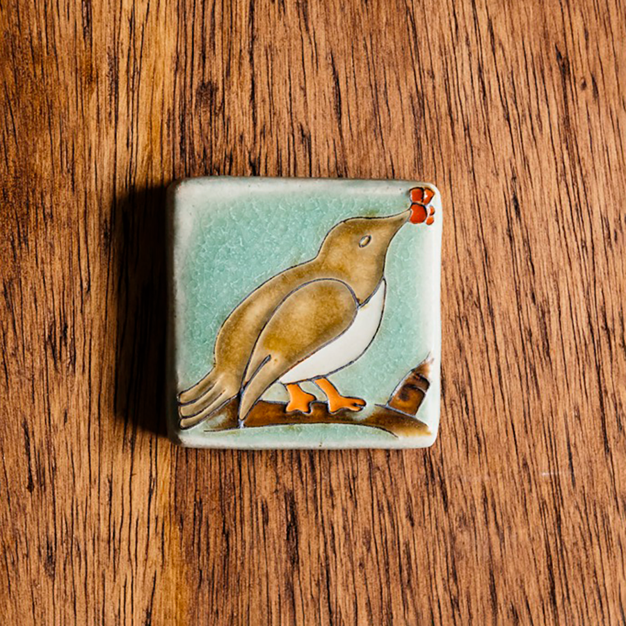 This Bird with Berries Tile depicts a brown bird sitting on a branch with red berries in its beak. This tile is hand-painted with brown, blue and red glazes.