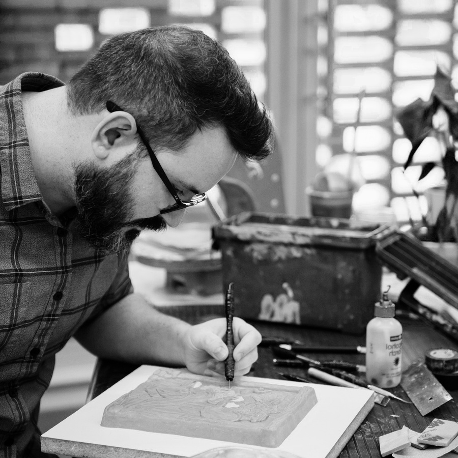 A Pewabic designer leans over his desk where he is using a small tool to intricately carve into an unfinished tile. 
