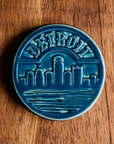 The Detroit Trivet features the Detroit skyline with the Renaissance Center in the middle. The word "Detroit" is carved along the top of the circular tile in an Olde English font similar to the one used for the Detroit Tigers' logo. This tile features the matte french blue Peacock glaze.