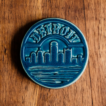 The Detroit Trivet features the Detroit skyline with the Renaissance Center in the middle. The word "Detroit" is carved along the top of the circular tile in an Olde English font similar to the one used for the Detroit Tigers' logo. This tile features the matte french blue Peacock glaze.