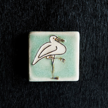 The Hand-Painted Crane Tile features a line drawing of a white crane with its whole body turned to the right, one leg on the ground and the other folded up, pointing to the right. The background is a pale greenish-blue.