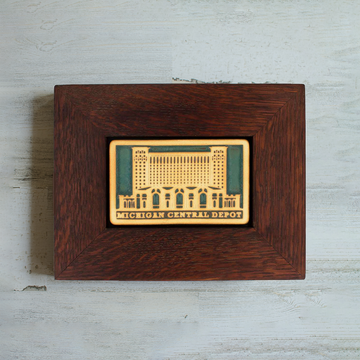 The Michigan Central Depot Tile features the image of the large many-windowed building with the words "Michigan Central Depot" across the bottom of the tile. The two toned nature of the tile means that the design and words have been scraped of their glaze giving them a creamy off-white look while the background of the image is in the crystalline, shimmering deep green Viridian glaze. The oak wood frame is a deep reddish brown color.