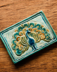 The Peacock Tile features a peacock facing straight ahead with its feathers fanned out showing off its colorful plumes. In the bottom right corner of the tile are the letters "M C S" in black writing. This is the only tile on which Pewabic's co-founder Mary Chase Stratton included a signature. The hand painted tile features bright blues, greens and beiges.