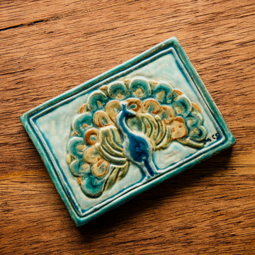 The Peacock Tile features a peacock facing straight ahead with its feathers fanned out showing off its colorful plumes. In the bottom right corner of the tile are the letters "M C S" in black writing. This is the only tile on which Pewabic's co-founder Mary Chase Stratton included a signature. The hand painted tile features bright blues, greens and beiges.